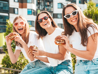 Three young beautiful smiling hipster female in trendy summer same clothes. Sexy carefree women posing in the street.Positive models having fun in sunglasses.Holding juicy burger and eating hamburger