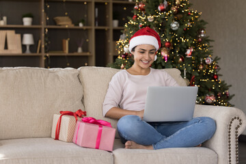 Full length smiling young beautiful Indian woman using computer, web surfing information, choosing Christmas movie online, reading wishes or congratulating friends with New Year in social network.