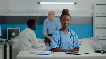 Portrait of nurse typing on laptop keyboard in medical office at facility. Assistant woman with uniform sitting at desk using gadget with technology. Doctor and old patient in background