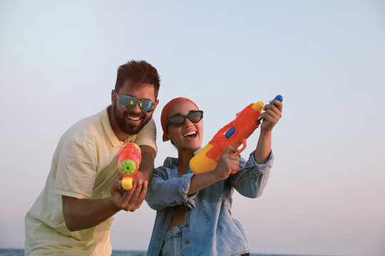 Happy couple with water guns against light blue sky at sunset
