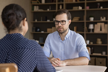 Concentrated young male applicant in eyewear holding job interview conversation with professional mixed race female hr manager, sitting at table in modern office, corporate recruitment concept.