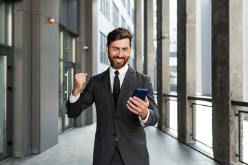 Successful and happy businessman shouts with joy and satisfaction of winning, reads good news from a mobile phone, outside office looks at camera