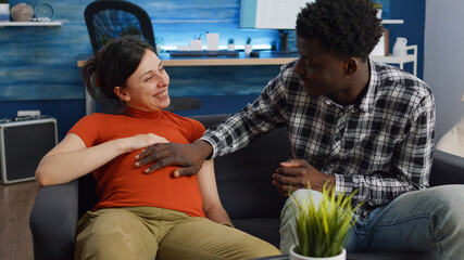Pregnant interracial couple sitting on sofa at home while black father of child holding hand on belly. Woman expecting baby and relaxing on couch. Multi ethnic family with pregnancy