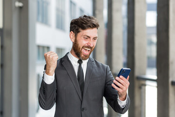 Successful and happy businessman shouts with joy and satisfaction of winning, reads good news from a mobile phone, outside office looks at camera