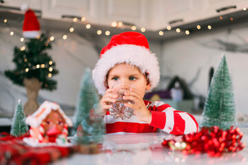 Fototapeta na wymiar Cute boy kid in a red Santa hat drinking filtered water from a glass in the kitchen. Holidays, health concept.