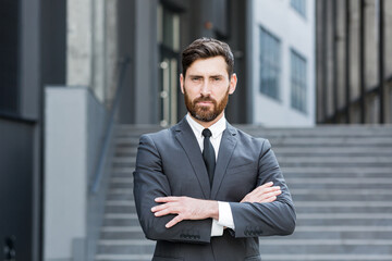 portrait young confident successful Caucasian bearded businessman looking at camera Serious business man standing on urban background of modern office building outdoors Male a formal suit arms crossed - obrazy, fototapety, plakaty