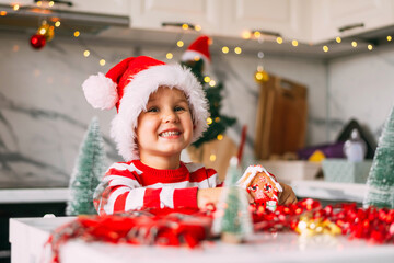 Fototapeta na wymiar Funny boy child 3 y.o. in a Santa hat is sitting in the kitchen with a decor for Christmas. Holidays, Traditions, baby food and health concept.