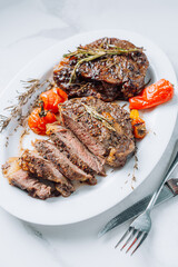 juicy well-fried ribeye steaks on a white platter on a white marble surface with raspmarine and fried cherry tomatoes and peppers. steaks are sliced and ready to serve