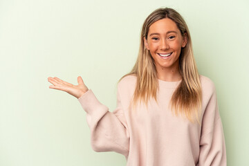 Young caucasian woman isolated on green background showing a copy space on a palm and holding another hand on waist.