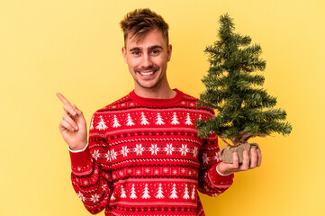Young caucasian man holding a Christmas tree isolated on yellow background smiling and pointing aside, showing something at blank space.
