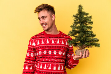 Young caucasian man holding a Christmas tree isolated on yellow background looks aside smiling, cheerful and pleasant.