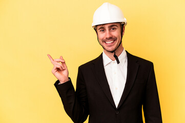 Young architect caucasian man isolated on yellow background smiling and pointing aside, showing something at blank space.