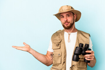 Young caucasian man looking at animals through binoculars isolated on blue background showing a copy space on a palm and holding another hand on waist.