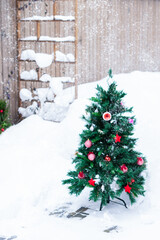 Artificial fir tree decorated with Christmas red toys outdoors during snowfall in yard in snowy background, New Year and Christmas concept