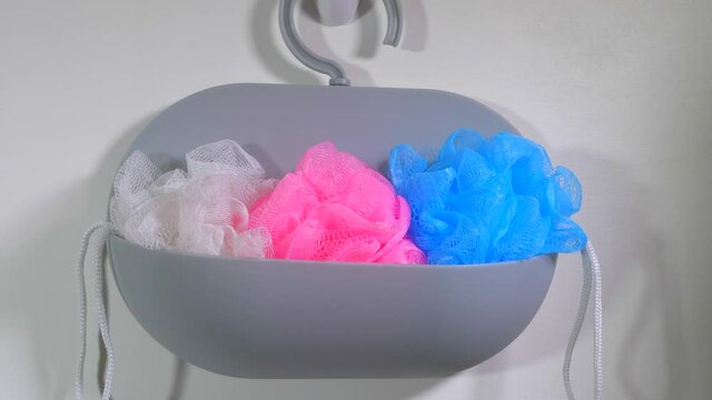 Closeup POV shot of a man&rsquo;s hand hanging on a wall, three new pink, blue and white bathroom exfoliating scrunchies / puffs pressed into a plastic holder.