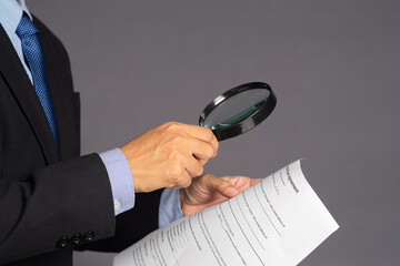 Businessman in a suit holding a magnifying glass and reading a document while standing over a gray...