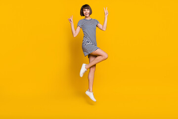 Full size photo of joyful millennial lady jump show v-sign wear striped dress sneakers isolated on yellow background