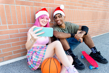 merry christmas! happy diverse couple of friends wearing xmas santa hat making a selfie portrait sitting against a background wall together on winter warm festivities outdoors. summer holidays concept - Powered by Adobe