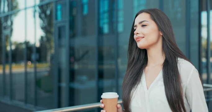 Beautiful cheerful woman working in an office, corporation, company, dressed in elegant clothes walks in front of a glass building, looks around smiling, coffee break, rest from her duties