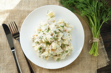 Traditional German potato salad with cucumber and mayonnaise. Top view. Potato salad with fresh radishes in a white bowl with dill and green onion on a rustic wooden table.