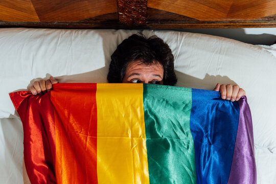 middle aged young man lying in bed covering himself with lgbtq flag feeling fear