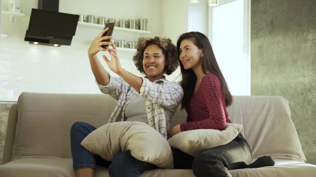 Happy young two girlfriends, an African-American and an Asian, take selfies in their house