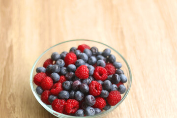 Glass bowl filled with raspberries and blueberries on wooden table. Selective focus.