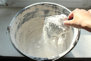 Close-up of a woman's hand with a spatula pouring dry putty into a bucket for kneading the solution. DIY home repair, interior reconstruction
