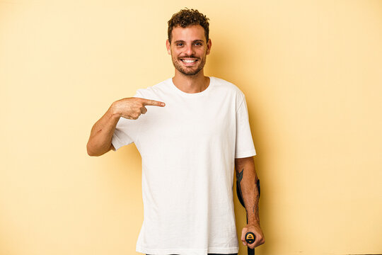 Young caucasian man holding crutch isolated on yellow background person pointing by hand to a shirt copy space, proud and confident
