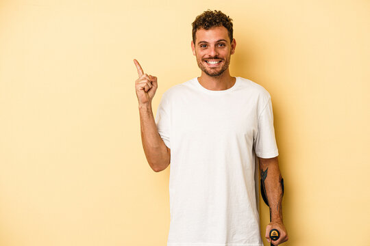 Young caucasian man holding crutch isolated on yellow background smiling and pointing aside, showing something at blank space.