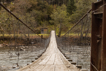 Old wooden suspension bridge across mountain river among autumn forest. Moody image. Countryside background.