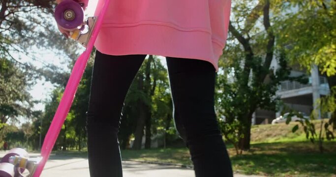Young woman in pink sweatshirt and black skinny trousers walks limping on park alley holding penny board in hand after skating on sunny day