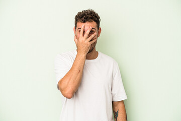 Young caucasian man isolated on green background blink at the camera through fingers, embarrassed...