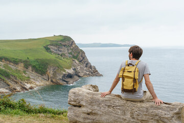 Adventurous caucasian man isolated sit on a cliff bench. Horizontal panoramic view of man with backpack traveling in the coast. People and travel concept.