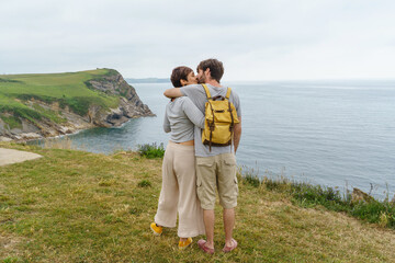 Unrecognizable couple in love kissing and watching the sea on a cliff. Horizontal view of backpackers sightseeing in holidays. Travel and people concept.