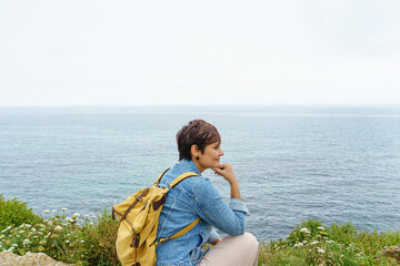 Cheerful caucasian woman isolated sit on a cliff bench. Horizontal view of woman traveling in the coast with blue sea in the background. People and travel concept.