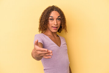 Young latin transsexual woman isolated on yellow background stretching hand at camera in greeting gesture. - obrazy, fototapety, plakaty