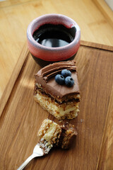 Piece of Kiev cake with chocolate icing and blueberries and cup of coffee on wooden board. A bite of cake on a fork