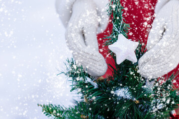 Woman’s hands with knitted white mittens gloves hanging Christmas toys on artificial fir tree branches during snowfall in winter time, copy space