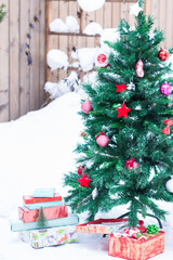 Colorful boxes of gifts on snow and artificial fir tree decorated with Christmas red toys outdoors in yard in snowy background, New Year and Christmas concept