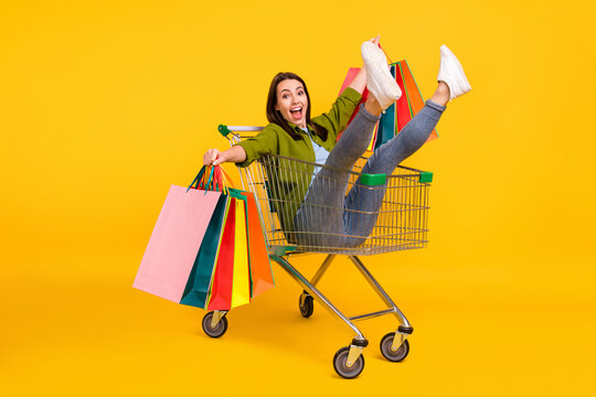 Photo of pretty funky young lady wear green shirt smiling holding shopping bags riding cart isolated yellow color background