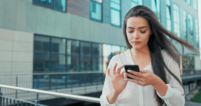 Attractive girl working in office corporation, company, dressed in elegant outfit, stands leaning against railing in front of glass building, holding a phone in hand, sending messages, chatting away
