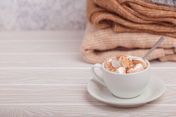 Cozy winter composition, cup with hot chocolate or cocoa with marshmallow and pile of warm knitted clothes on white wooden rustic background. Comfy and soft winter at home, free space for text