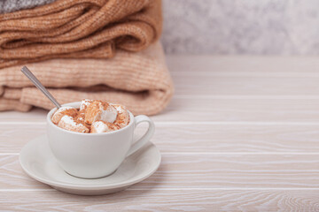 Cozy winter composition, cup with hot chocolate or cocoa with marshmallow and pile of warm knitted clothes on white wooden rustic background. Comfy and soft winter at home, free space for text