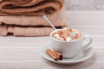 Cozy winter composition, cup with hot chocolate or cocoa with marshmallow and pile of warm knitted clothes on white wooden rustic background. Comfy and soft winter at home, free space for text