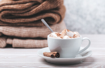 Cozy winter composition, cup with hot chocolate or cocoa with marshmallow and pile of warm knitted clothes on white wooden rustic background. Comfy and soft winter at home, free space for text
