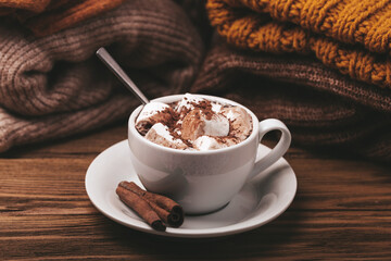 Cozy winter composition, cup with hot chocolate or cocoa with marshmallow and pile of warm knitted clothes on wooden rustic background. Comfy and soft winter at home, winter or autumn concept