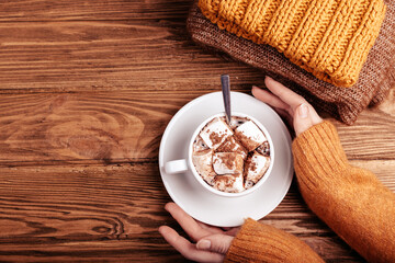 Fototapeta na wymiar Cozy winter flat lay, female hands holding cup with hot chocolate or cocoa with marshmallow and pile of warm knitted clothes on wooden rustic background. Comfy and soft winter or autumn concept
