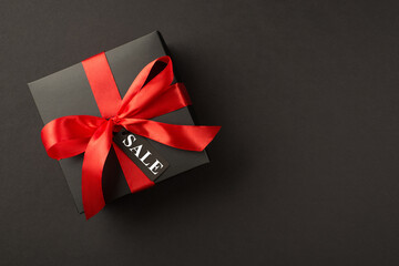 Top view photo of stylish giftbox in black packaging with vivid red satin ribbon bow and tag on isolated black background with text sale on pricetag