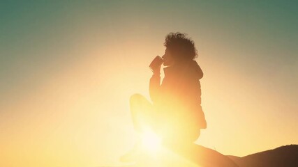 Rear view of woman hiker sitting on rock on top of hill while looking at sunset over mountain valley, relaxing drinking energy drink